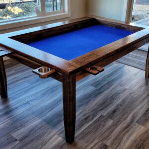 A table with a sunken middle surface for playing board games. sits in a living room with windows behind it, The table has cupholders and trays around the perimeter for holding drinks and game pieces